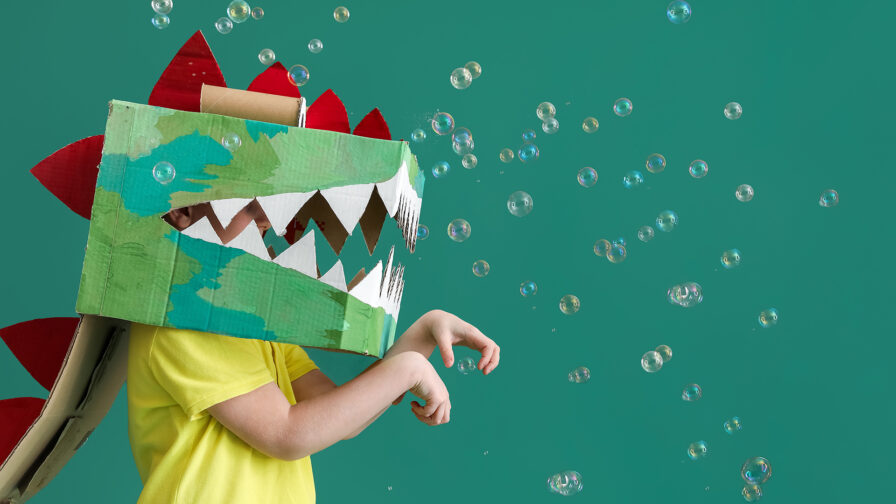 Little boy in cardboard dinosaur costume and soap bubbles on gre
